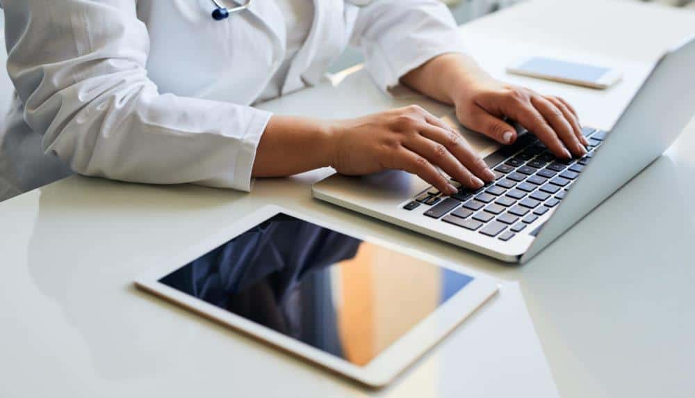 A Doctor is Typing on a Laptop Next to a Tablet