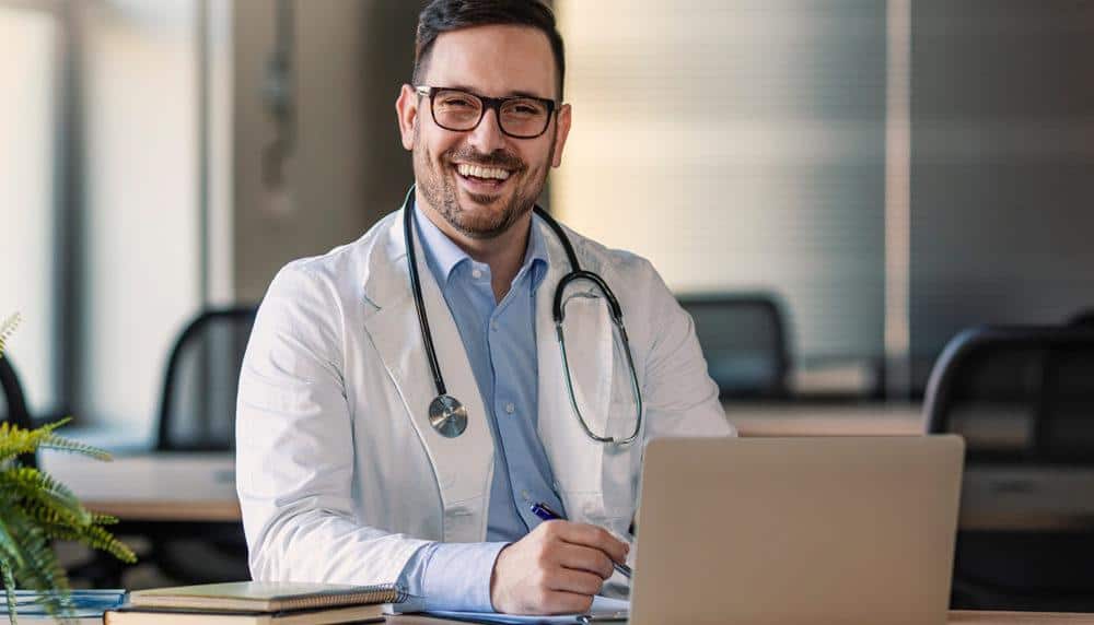 a smiling doctor is sitting at a desk with a laptop computer